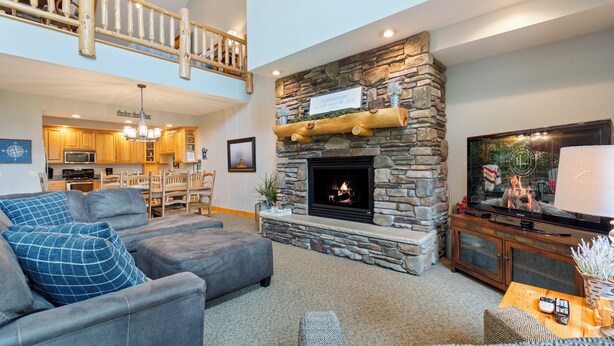 A spacious living room with a stone fireplace, a television, and a high ceiling with a wooden staircase.