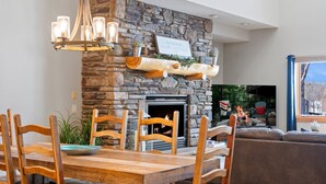 A dining area with a stone fireplace, wooden chairs, a wooden table, and a chandelier.