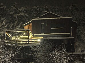 A wooden cabin with a balcony and a lit interior, surrounded by snow-covered trees.