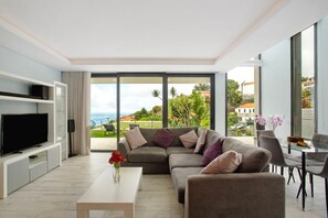 A modern living room with a large flat-screen TV, a sofa, a coffee table with a vase of flowers, and a balcony with a view of the sea and buildings. - Villa 'Jolie' with Sea View, Wi-Fi and Air Conditioning (Ponta do Sol)