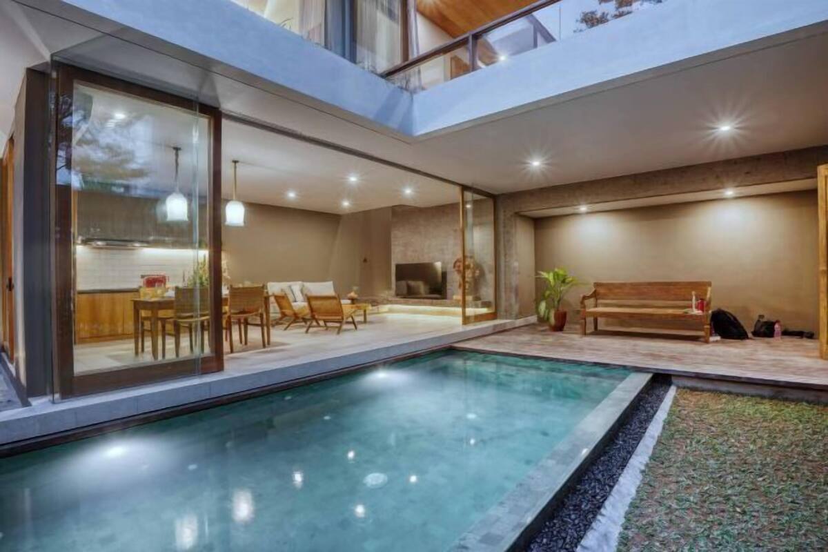 Una piscina interior moderna con paredes de vidrio, un banco de madera y un comedor con sillas.