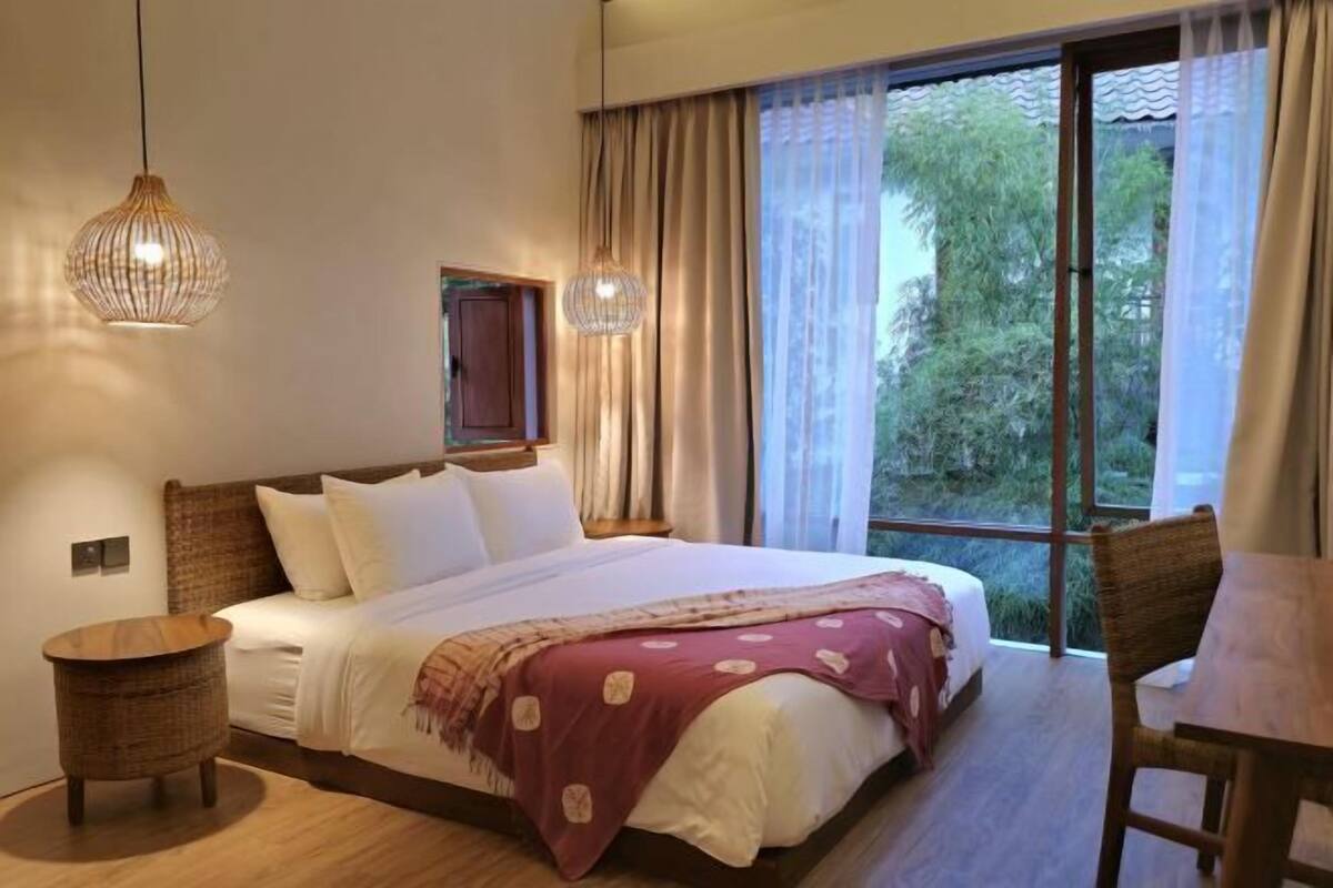 Un dormitorio con una cama grande, dos lámparas colgantes, una ventana con cortinas y vistas a la vegetación.