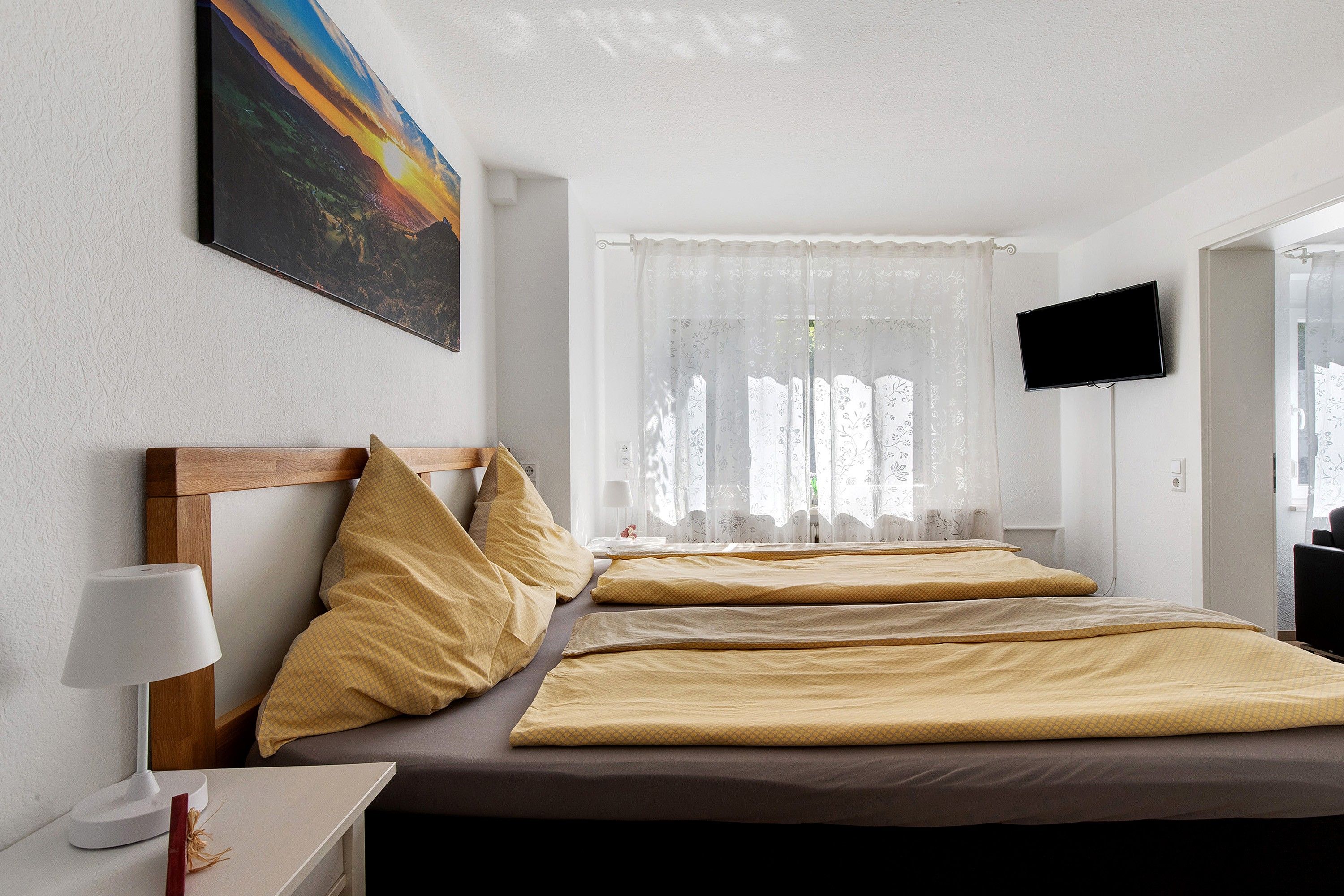 Et hotelværelse med en seng, et sengebord med en lampe, et fjernsyn monteret på væggen og et vindue med lette gardiner.