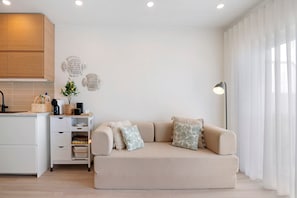 Un salon moderne avec un canapé beige, une petite table d’appoint et un coin cuisine doté de placards et d’un plan de travail.