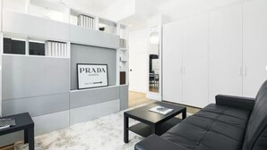 A modern living room with a black sofa, a coffee table, and a grey shelving unit with a Prada sign. - Acquileia - Stylish loft  steps from Corso Magenta (Milano)