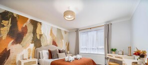 Ein modernes Schlafzimmer mit einem großen, abstrakten Gemälde, einem Bett mit vielen Kissen, einem Nachttisch mit einer Lampe sowie einem Schreibtisch mit Pflanze und Vase.