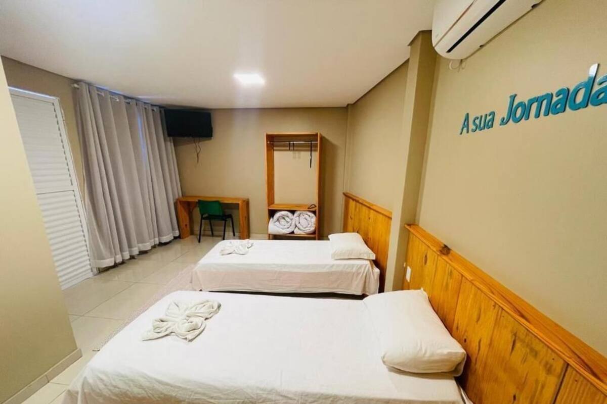 A hotel room with two beds, a wooden headboard, a flat-screen TV, a mirror, and a wall sign reading "Asua Jomada".