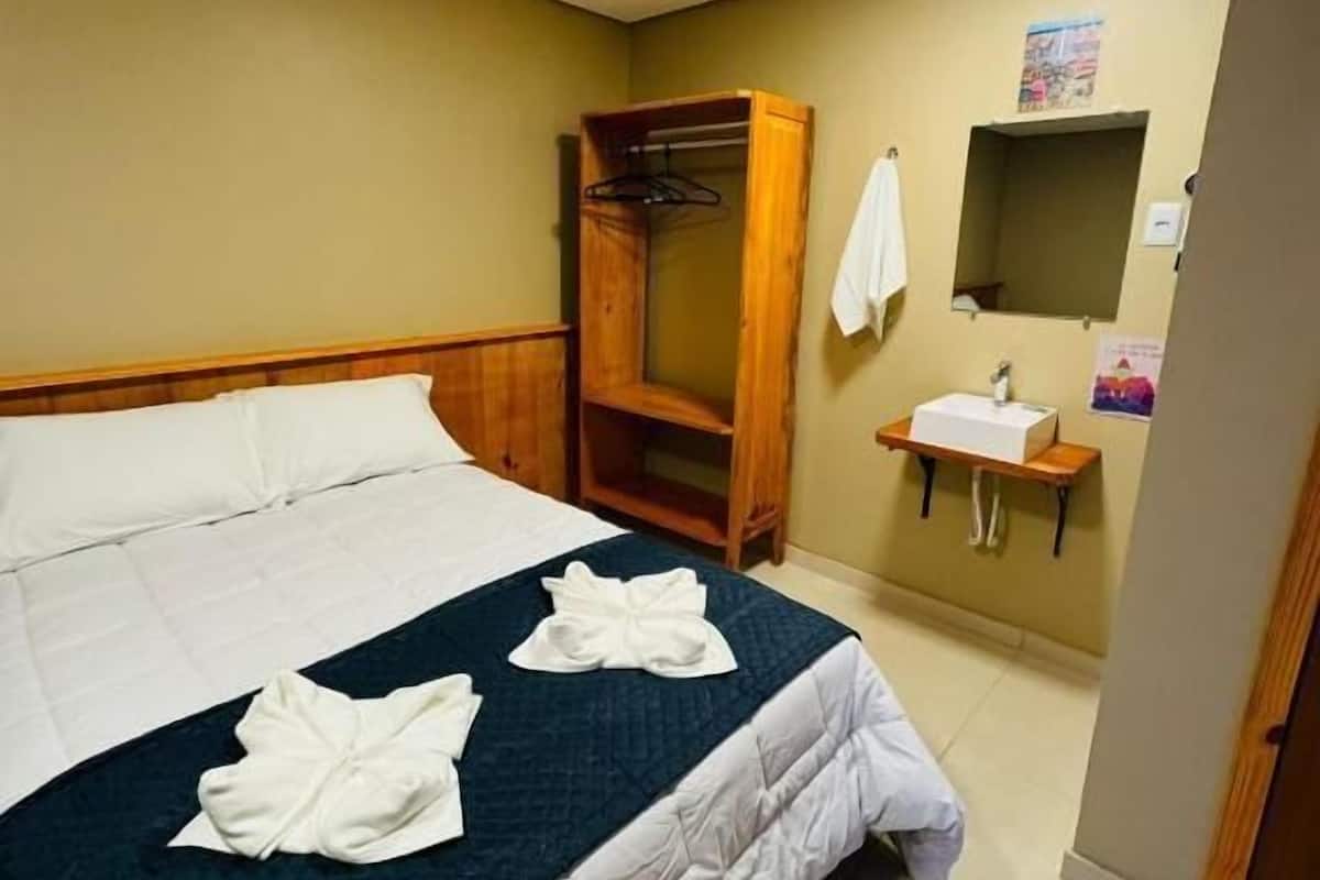 A hotel room with a bed, a wooden wardrobe, a small bathroom with a sink and mirror, and a poster on the wall.