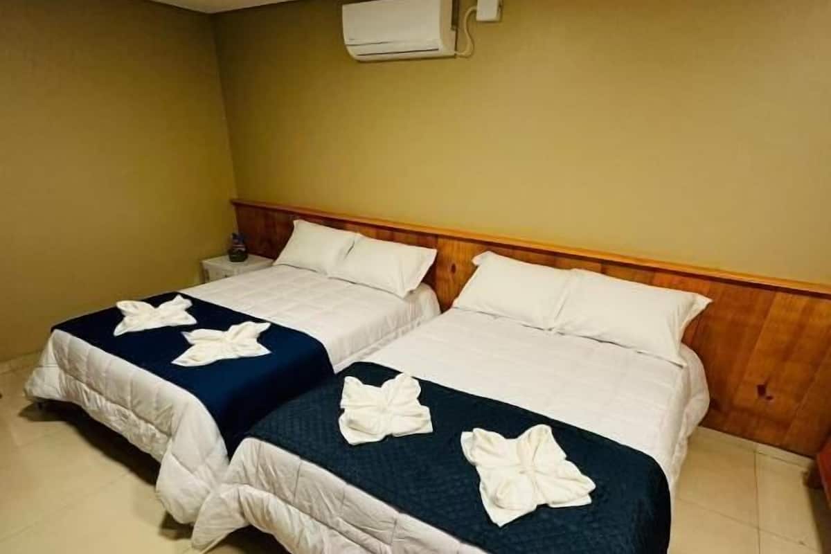 A hotel room with two beds, each with white bedding and decorative bows, a wooden headboard, and an air conditioning unit on the wall.