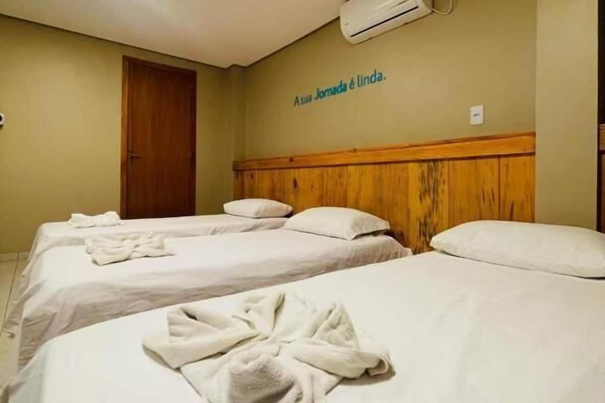 A hotel room with three beds, each with a towel on the bed. There is a wooden headboard and a wall-mounted air conditioner.