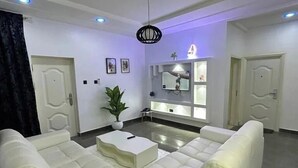 Una sala moderna con un sofá sectional blanco, un televisor de pantalla plana montado en la pared y una lámpara en el techo.