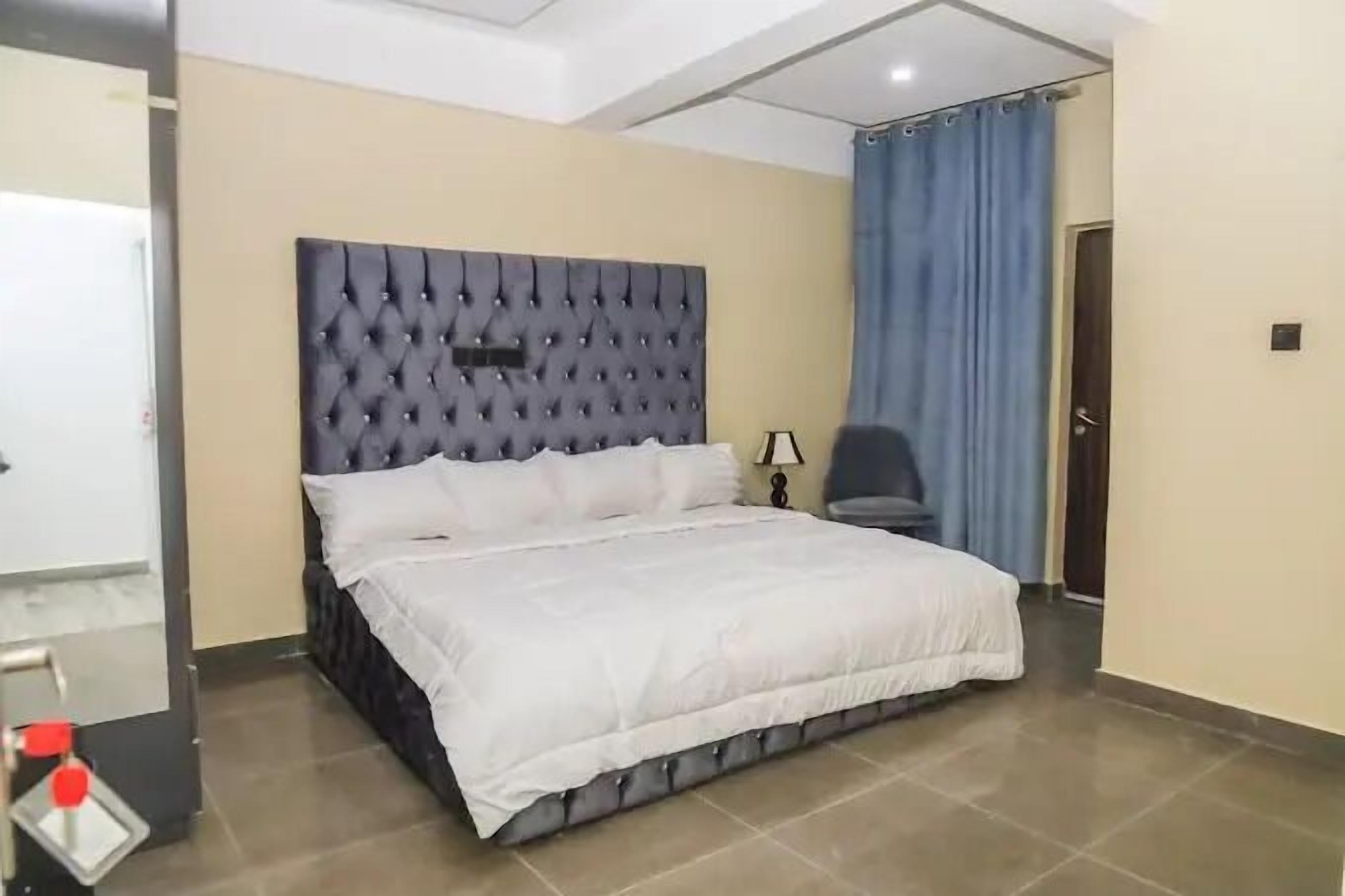 A bedroom with a tufted headboard, a large bed with white bedding, a nightstand with a lamp, and a blue curtain.