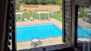 Vue depuis une chambre dotée d’une grande fenêtre donnant sur une piscine, un espace de détente extérieur et une colline lointaine sur laquelle se trouve une construction.