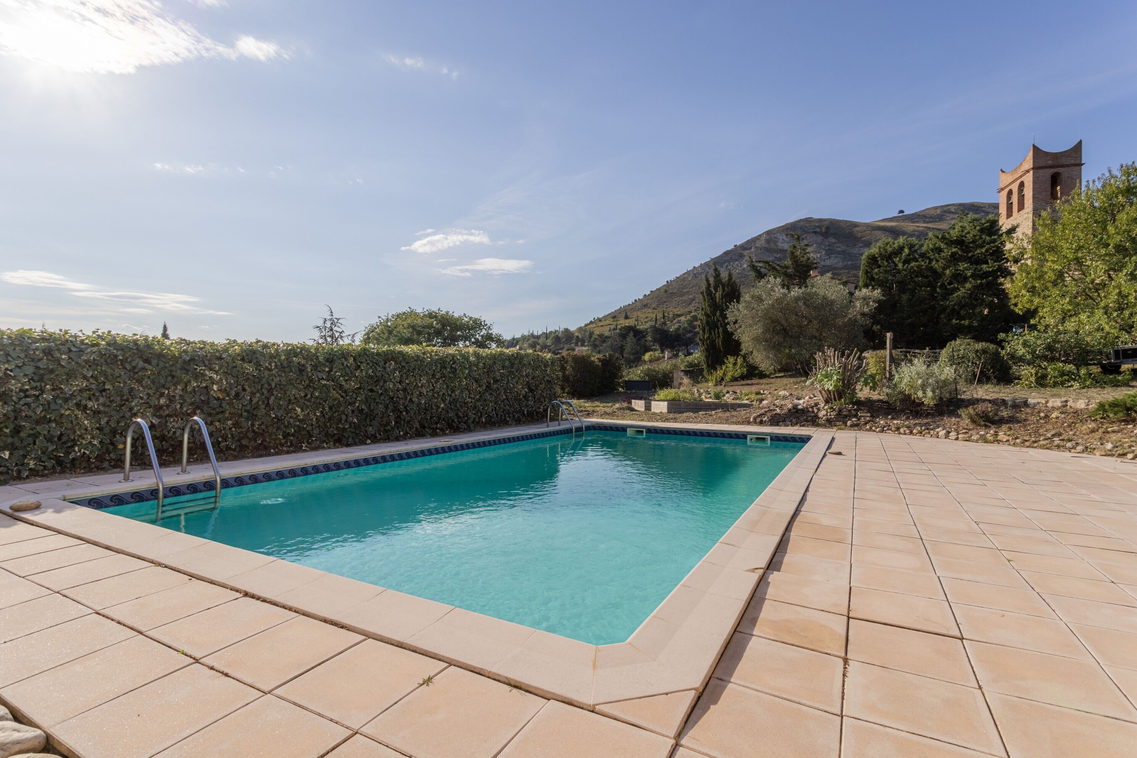 Une piscine rectangulaire, bordée d’une terrasse carrelée, entourée d’une haie et avec une montagne en toile de fond.