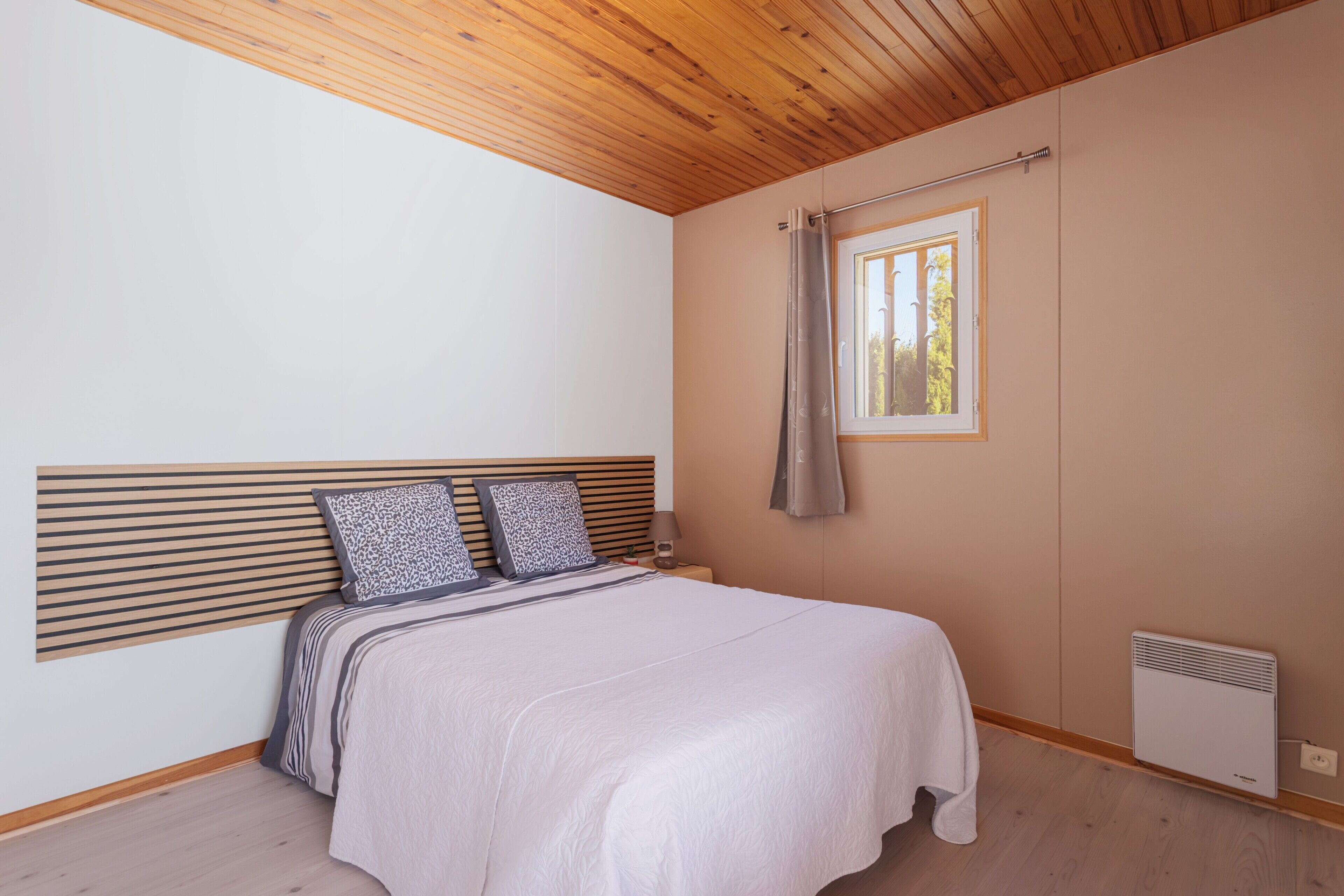 Quarto com teto de madeira, cama com cabeceira, janela com cortina e aquecedor fixado na parede.