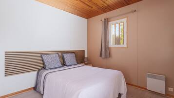 Quarto com teto de madeira, cama com cabeceira, janela com cortina e aquecedor fixado na parede.