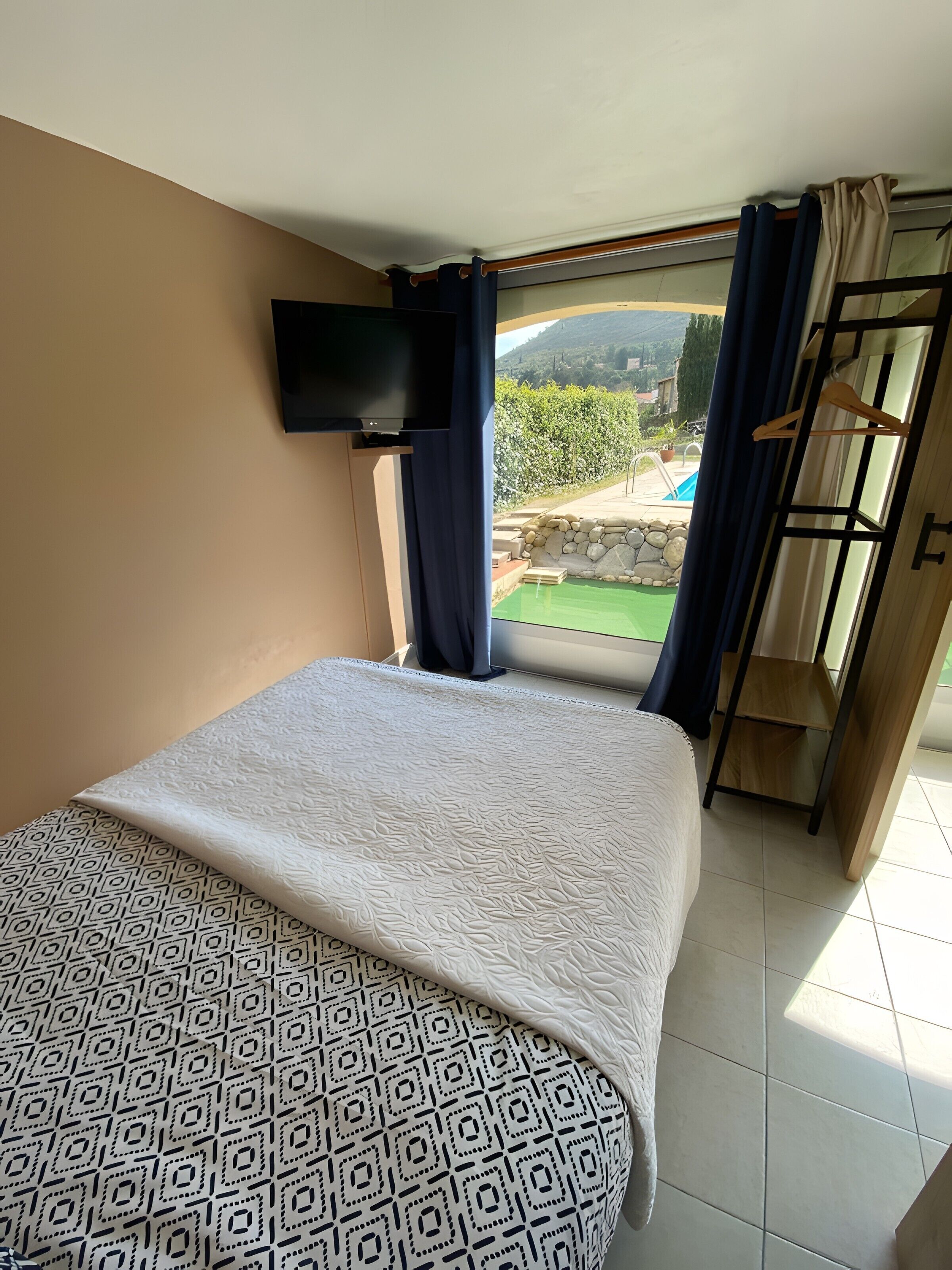 A bedroom with a bed, a TV mounted on the wall, a window with curtains, and a view of an outdoor swimming pool.