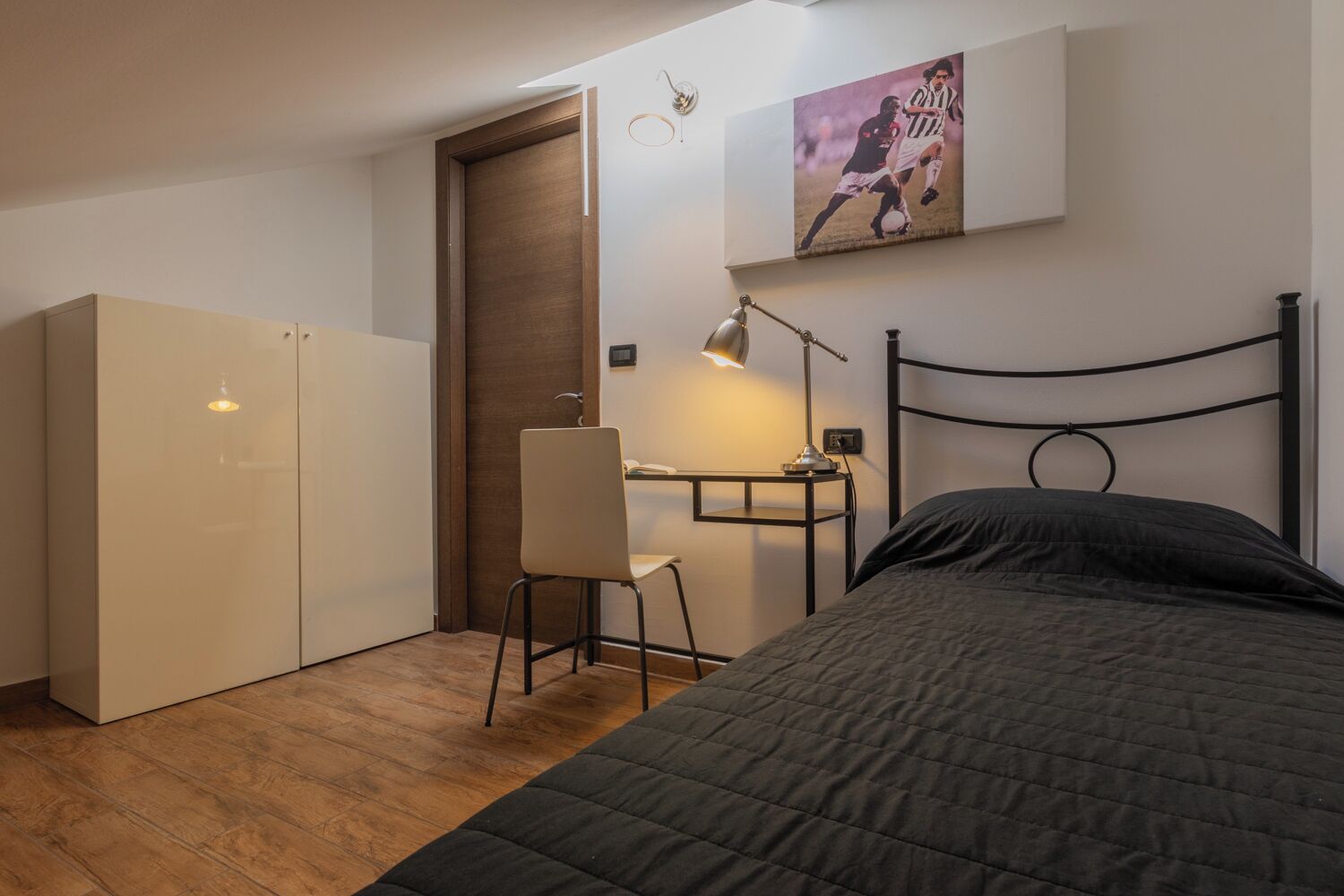 A compact bedroom with a bed, a desk with a chair, a wardrobe, and a wall-mounted picture.