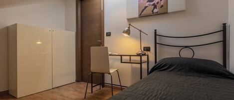 A compact bedroom with a bed, a desk with a chair, a wardrobe, and a wall-mounted picture.