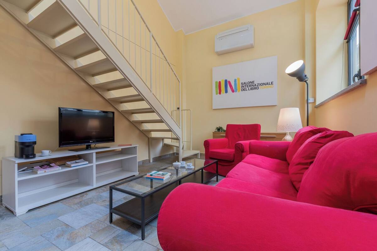 Un soggiorno con una scala, una TV a schermo piatto, divani rossi, una macchina da caffè e una libreria.