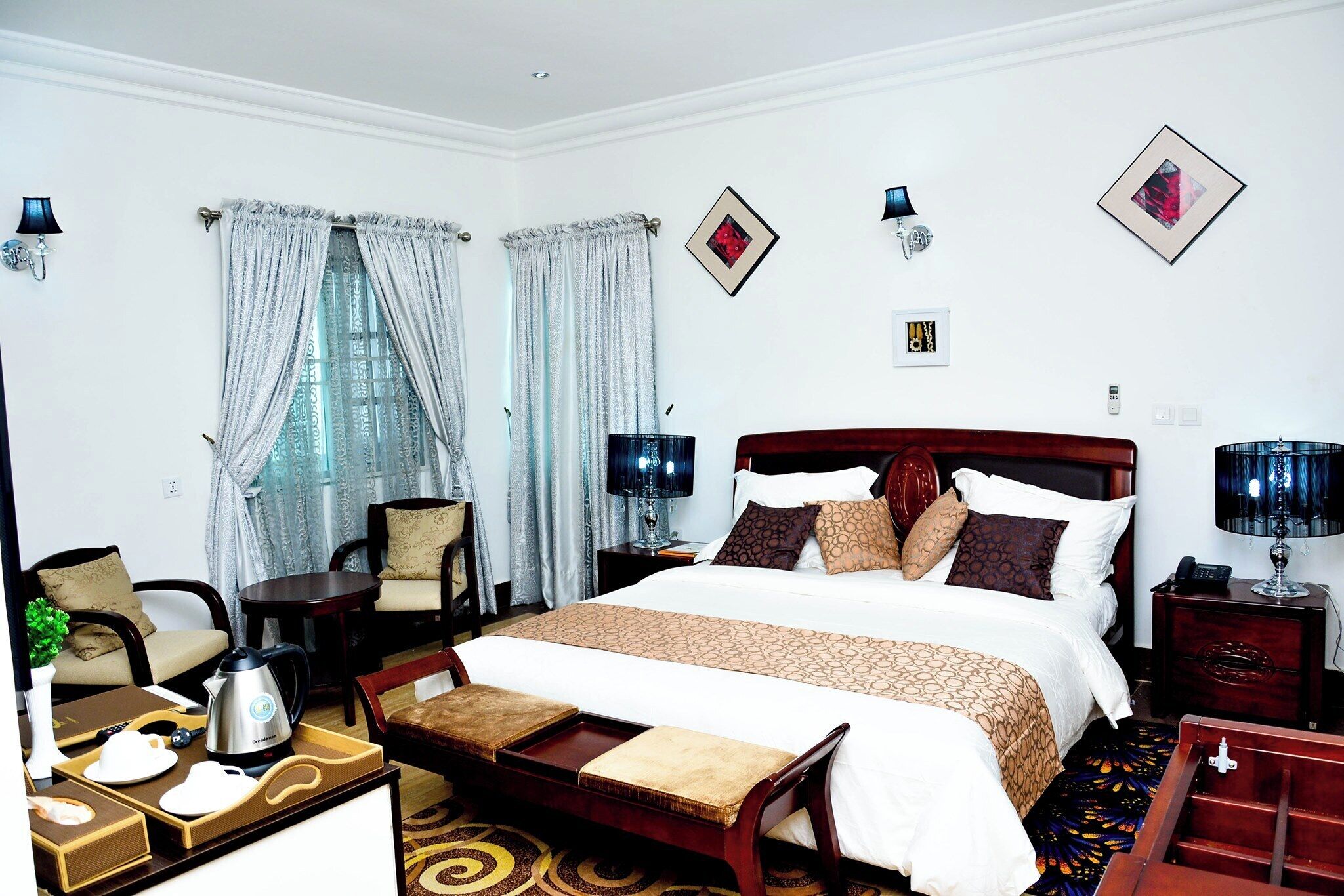 A hotel room with a large bed, two bedside tables with lamps, a wooden bench, a small table with a kettle, and a chair.