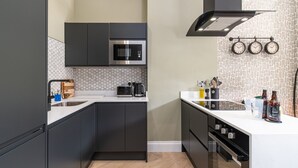 Uma cozinha moderna com armários escuros, micro-ondas, chaleira e máquina de café. Há três relógios na parede e uma pia com torneira.