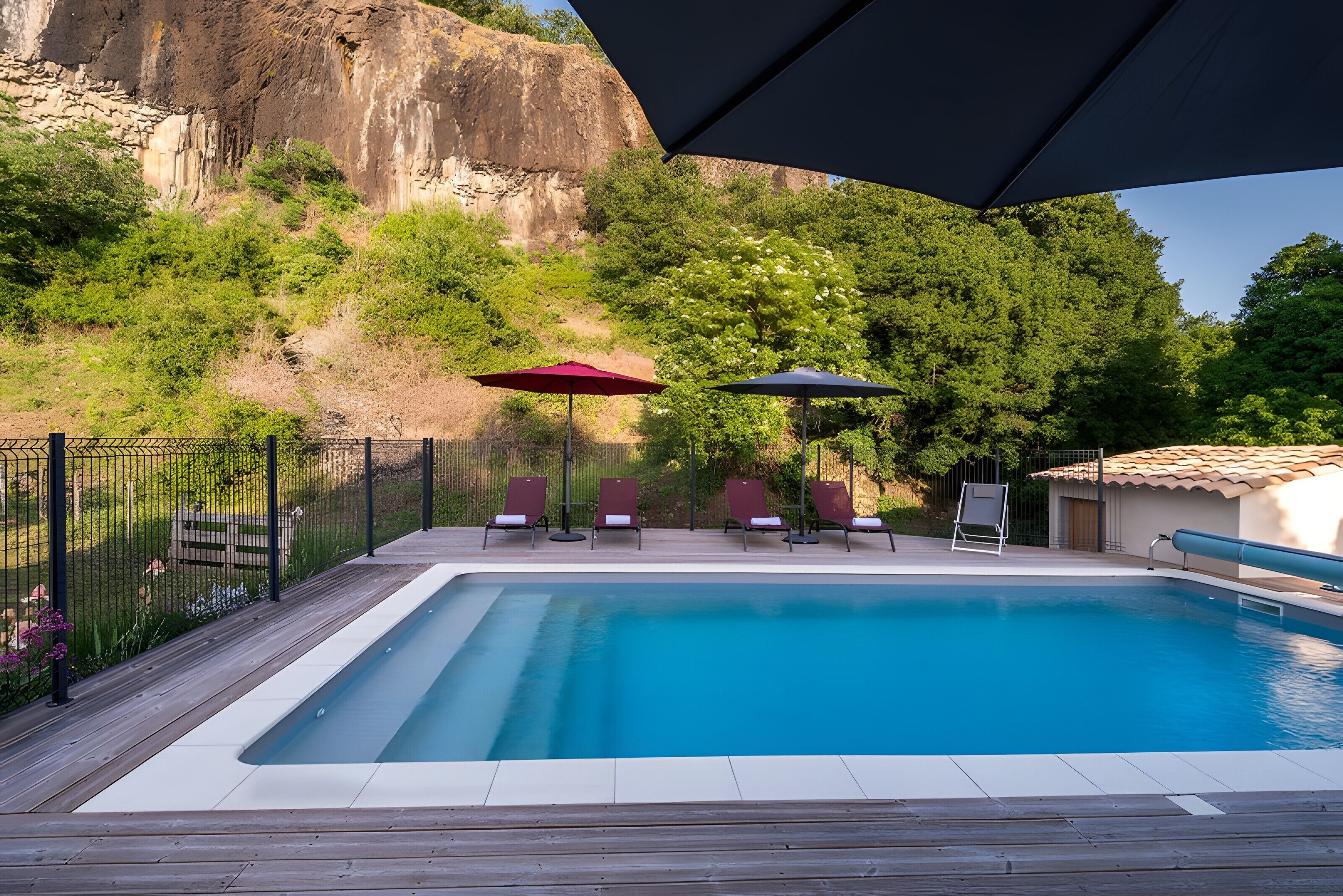 Une piscine entourée de chaises longues et de parasols, bordée d’une terrasse en bois et d’une zone délimitée par une clôture, avec une falaise rocheuse en arrière-plan.