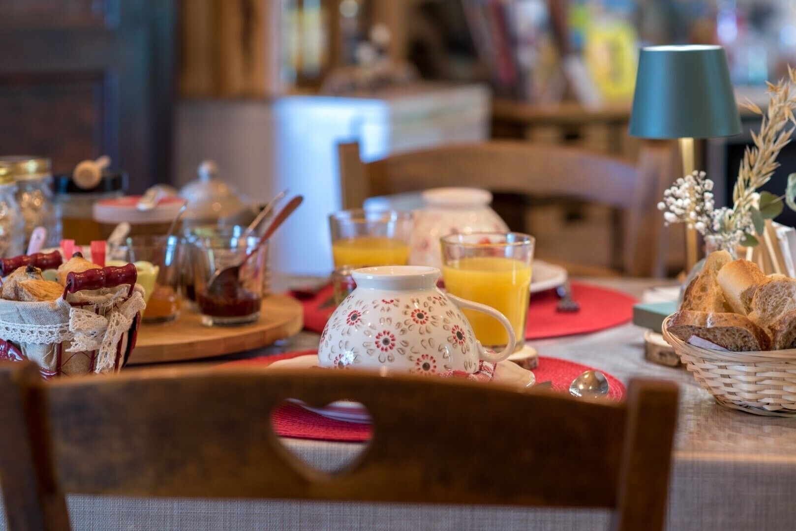 A breakfast setting with a table set for two, including a pitcher, glasses of orange juice, bread, and a basket of pastries.