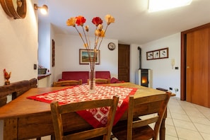 A dining area with a wooden table, chairs, a vase with flowers, a red tablecloth, a fireplace, and a clock on the wall.