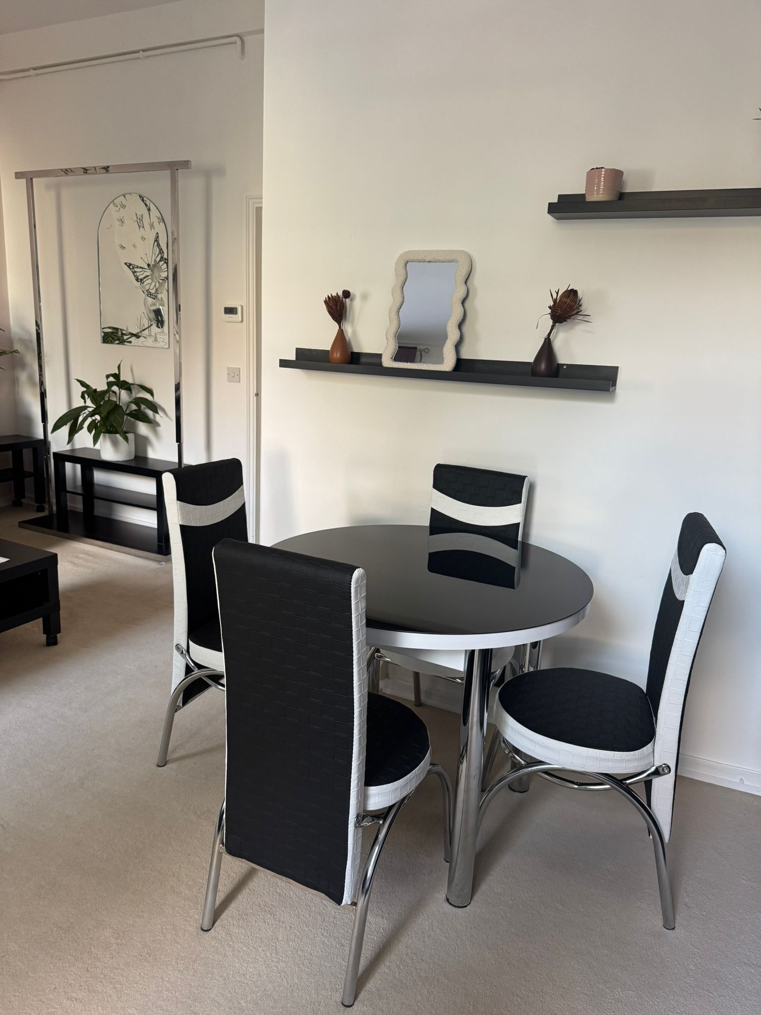 A dining area with a round table and four chairs, a small shelf with decorative items, and a door with a butterfly painting.