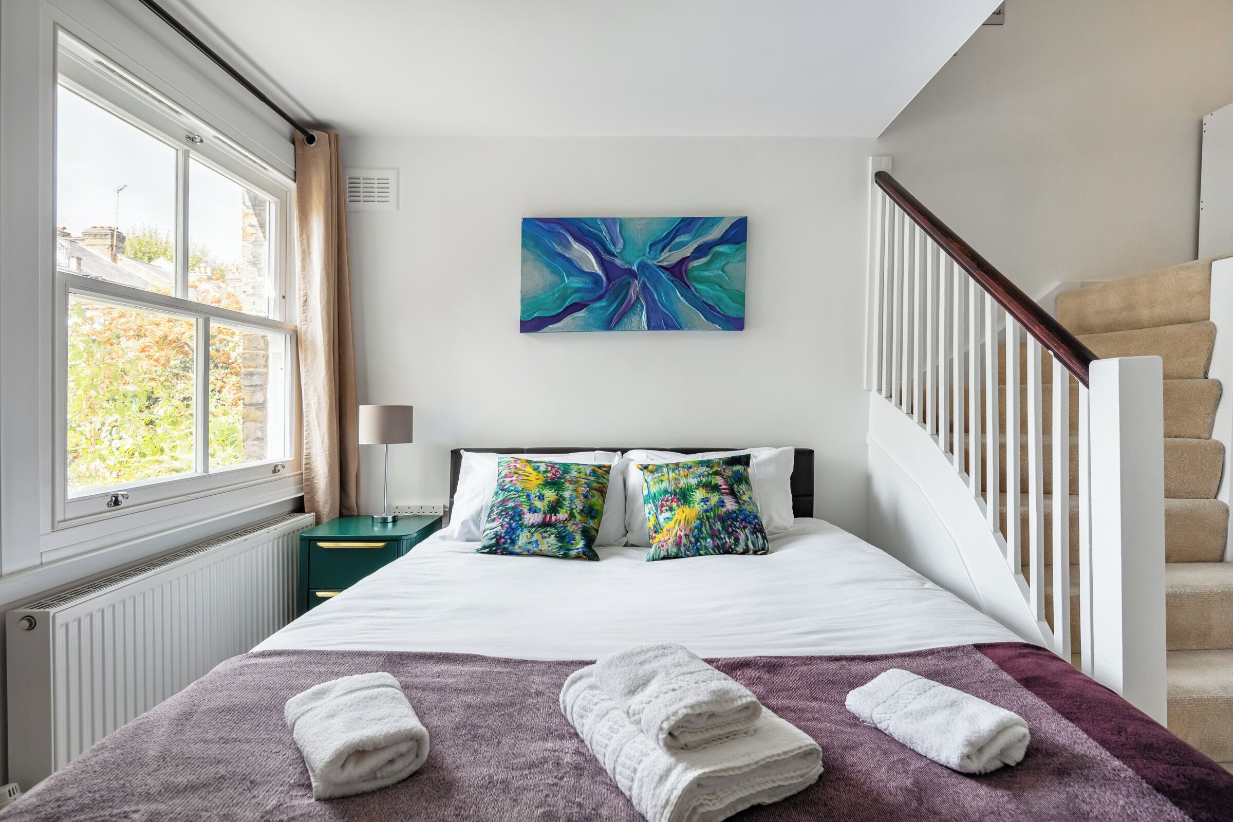 Ein Hotelzimmer mit einem großen Bett, zwei farbenfrohen Kissen, einer violetten Tagesdecke und Blick ins Freie durch ein Fenster.