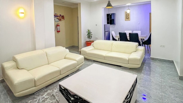 A modern living room with two cream-colored sofas, a white coffee table, and a dining area with chairs and a table. - Kpees Apartment (Abuja)