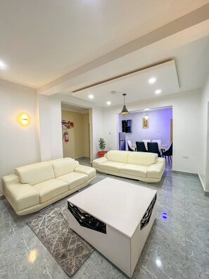 A modern living room with two cream-colored sofas, a white coffee table, and a dining area with chairs and a table. - Kpees Apartment (Abuja)