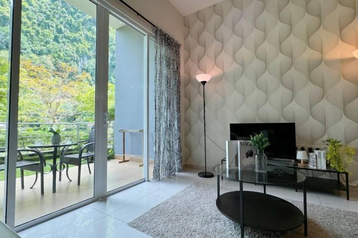 A modern living room with a large sliding glass door leading to a balcony, a flat-screen TV, a glass coffee table, and a patterned wall.