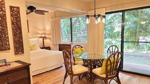 A hotel room with a bed, a wooden dresser, a glass table with chairs, and large windows with curtains.