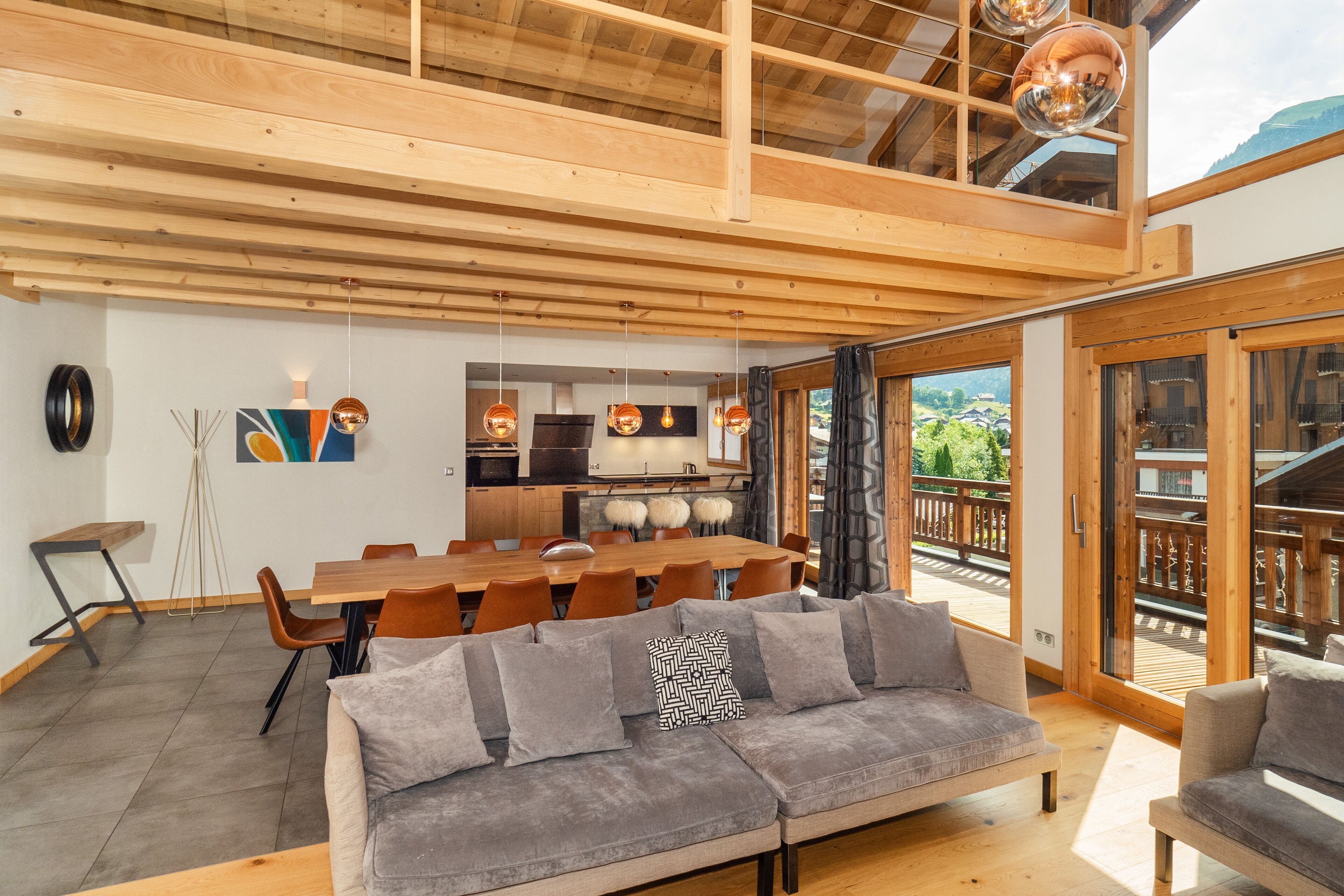 Un interior moderno con techo de madera, una zona de comedor con una mesa grande y sillas, un sofá con cojines y vistas al exterior a través de amplios ventanales.