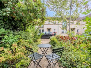 Een tuin met een tafel en stoelen, omgeven door weelderige begroeiing en een gebouw op de achtergrond.