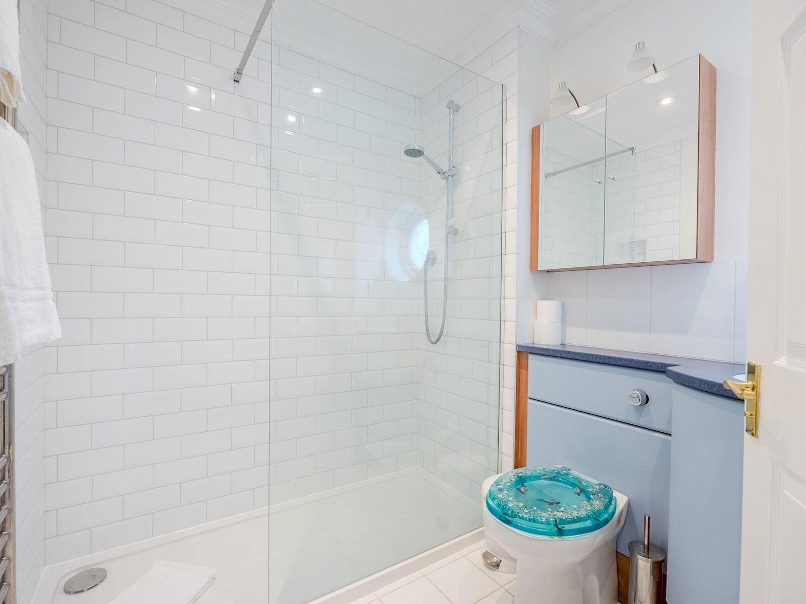A modern bathroom with white tiled walls, a glass shower enclosure, a toilet with a decorative blue lid, and a vanity with a mirror and a towel hanging on the wall.