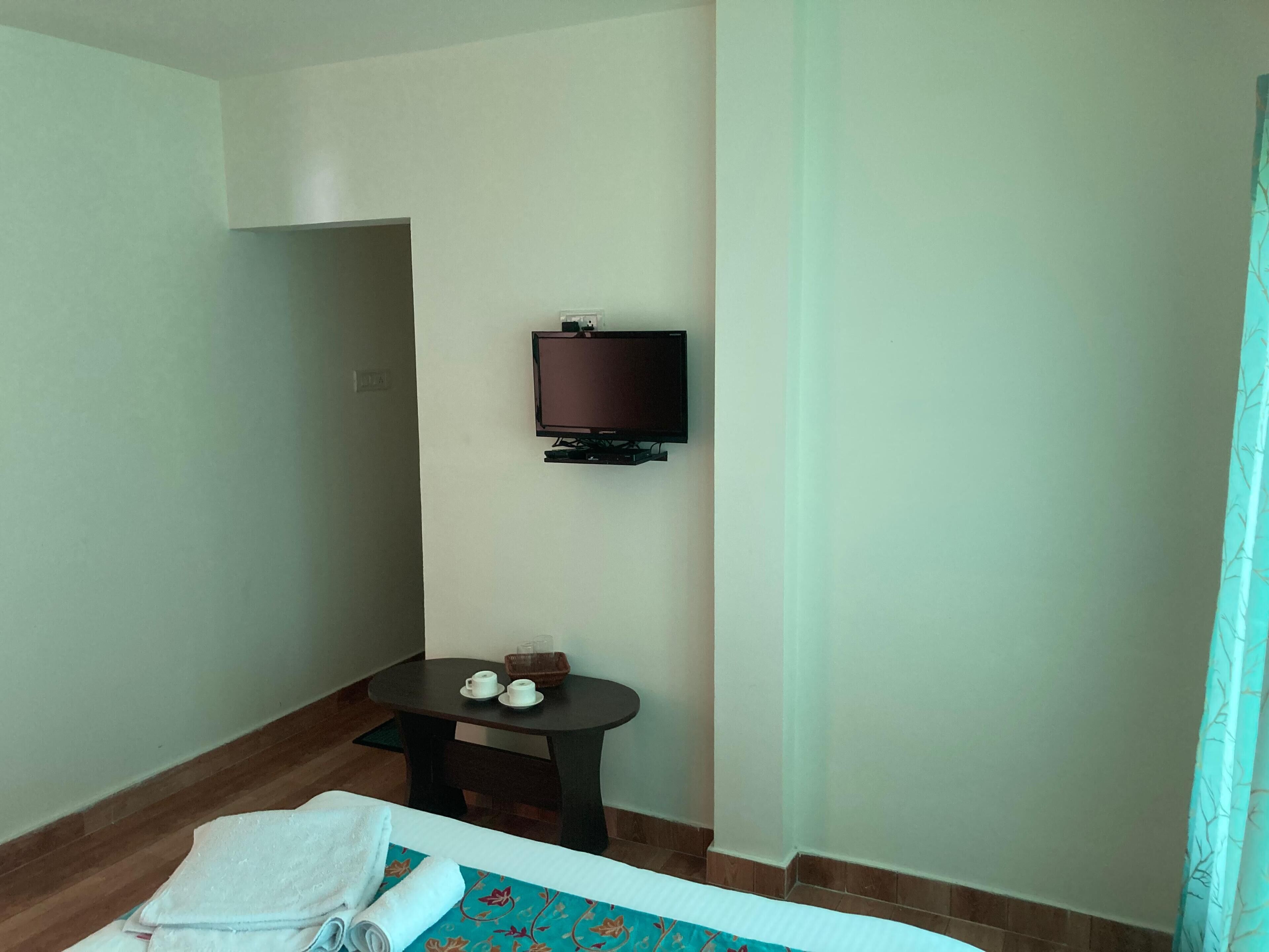 A hotel room with a bed, a small table with cups and a bowl, a television on the wall, and a window with a curtain.