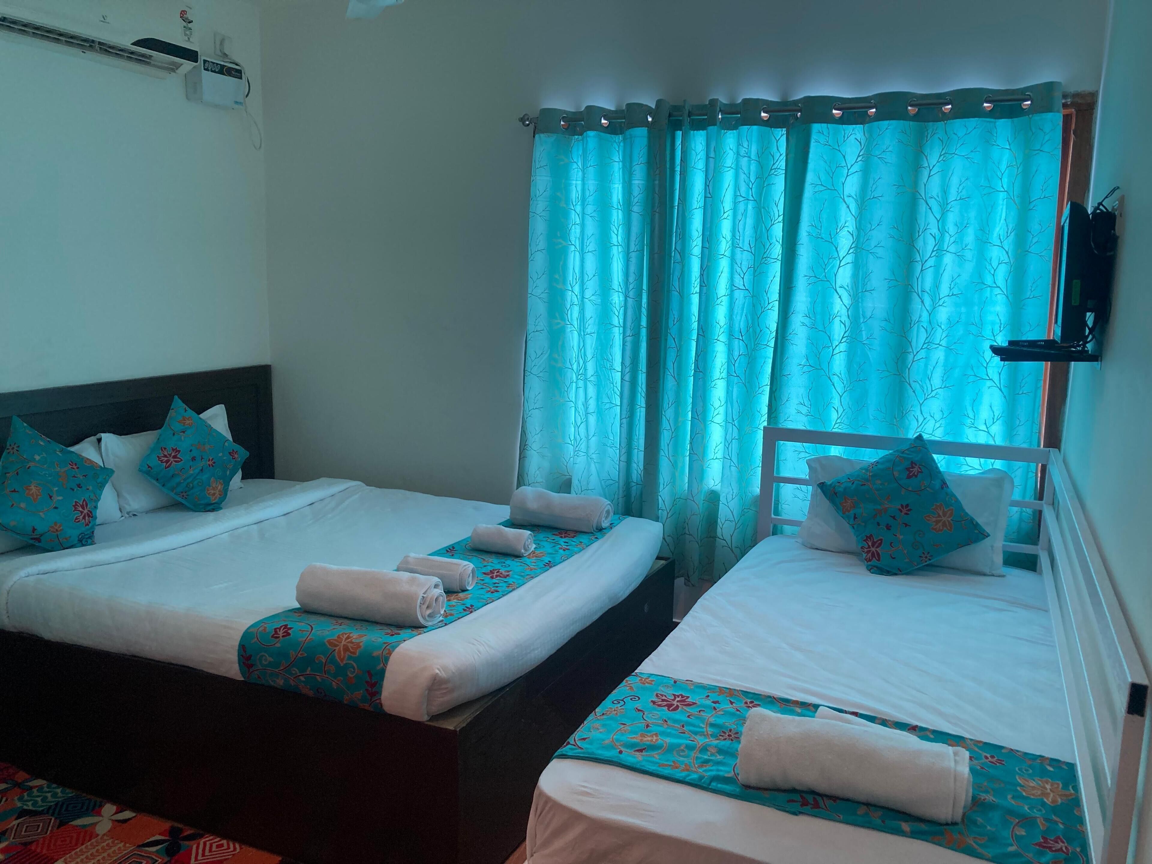 Camera d'albergo con due letti, ognuno con copripiumino bianco e cuscini coordinati, e un corridino da letto a motivi teal. Si vede una finestra con tende teal e un condizionatore d'aria a muro.