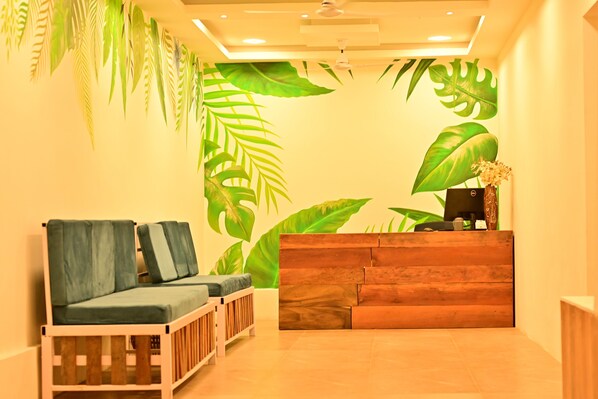A reception area with a wooden reception desk, two cushioned seating areas, and large tropical leaf wall art. - Rest Nest - Havelock (Port Blair)