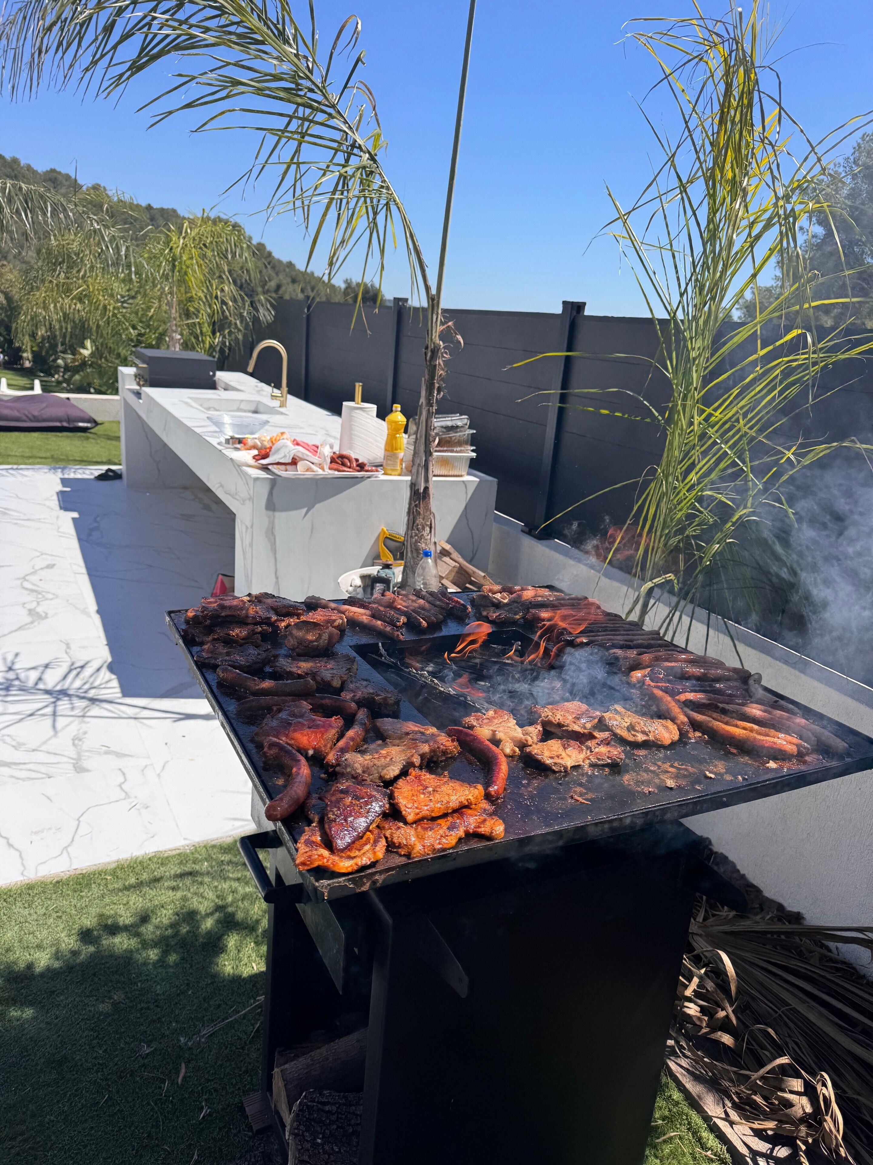 An outdoor barbecue grill with various meats being cooked, a sink, and a bottle of sauce.