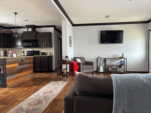 A modern kitchen with dark cabinetry and a wooden island, a living area with a gray sofa and a red chair, and a flat-screen TV mounted on the wall. - Spacious 3BR Rural Home + Barn Theater One Hour from Dallas, Fort Worth & Waco (Rice)