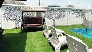 A backyard with artificial grass, a swing set, a pool, and a mural of trees.