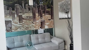 A modern living room with a grey sofa, a glass coffee table, a painting of a tree, and a large mural of ancient ruins.