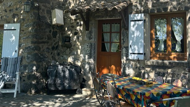 A stone building with a wooden door, windows with shutters, a table with chairs, and a covered outdoor seating area.