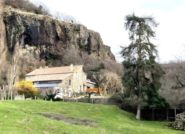 A stone house with a tiled roof, surrounded by a grassy area and a cliff.