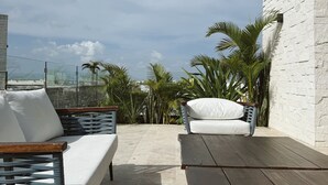 A rooftop terrace with two cushioned seating areas, a wooden table, and a white brick wall.