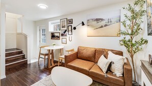 Een woonkamer met een bruine leren bank, een witte salontafel, een eettafel met stoelen en een trap naar een andere kamer.