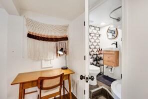 Una stanza con una scrivania e una sedia in legno, un arazzo macramè e un bagno con il water, il lavandino e lo specchio.